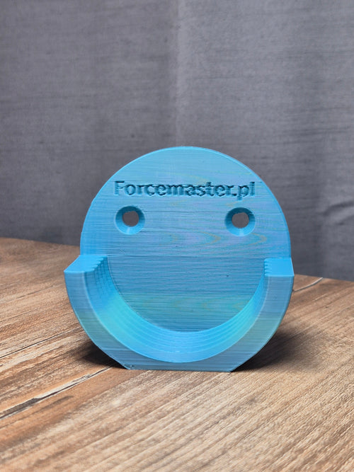 Niebieski element 3D z plastiku, uśmiechnięta buźka, grawerowany napis Forcemaster.pl, na drewnianym stole